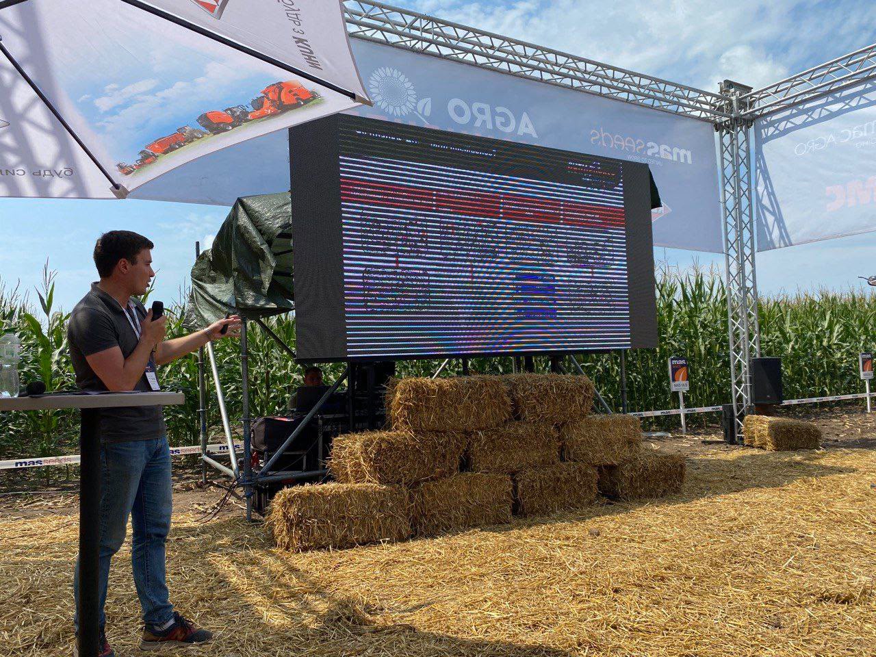 An outdoor agricultural technology presentation with a large screen and hay bales